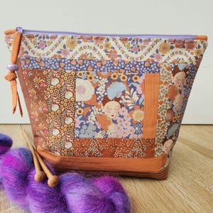 Peut inclure: Une pochette zippée en tissu patchwork avec des motifs floraux et des visages stylisés dans des tons orange, marron, violet et crème. Une fermeture éclair violette se trouve en haut, et une pelote de laine violette avec des aiguilles en bois est posée devant.