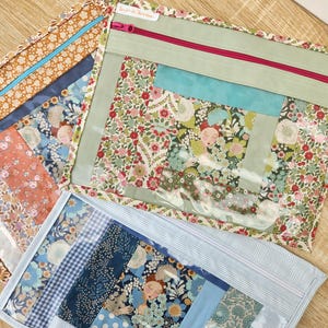 Peut inclure: Trois pochettes en tissu avec des fenêtres en vinyle transparent et des fermetures éclair. Les pochettes présentent des tissus à motifs floraux dans les tons de bleu, orange et vert. Une pochette a une fermeture éclair rose, et une autre a une fermeture éclair bleue.
