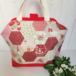 Peut inclure: Sac fourre-tout fait main avec un motif patchwork hexagonal rouge, crème et blanc. Le sac a une base et un dessus rouges unis, avec des anses crème. Motifs floraux et de Noël ornent le patchwork.