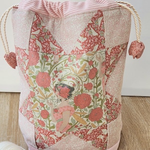 Peut inclure: Une pochette à cordon avec un motif patchwork composé de fleurs et de plantes dans des tons rose, vert et crème. Le panneau central représente une silhouette entourée de fleurs et de libellules. Le haut et la base sont en velours côtelé rose.