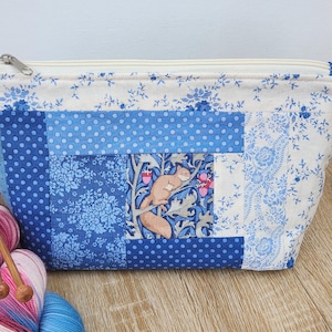 Peut inclure: Une trousse à fermeture éclair en tissu rectangulaire avec un motif patchwork de fleurs bleues et blanches, des pois, et un panneau central carré représentant un écureuil brun sur une branche avec des glands roses sur fond de feuillage bleu.
