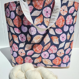 Peut inclure: Un sac fourre-tout bleu marine orné d'un motif répétitif de divers coquillages dans des tons orange, rose et blanc, avec deux anses en tissu blanc cassé. Un écheveau de fil torsadé épais de couleur crème est posé devant le sac.