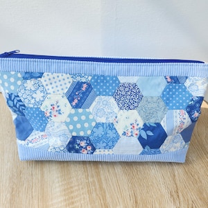 Peut inclure: Une trousse rectangulaire à fermeture éclair composée d'un patchwork de pièces de tissu hexagonales bleues et blanches avec divers motifs floraux, à pois et rayés. Les bords sont garnis de tissu velours côtelé bleu clair.