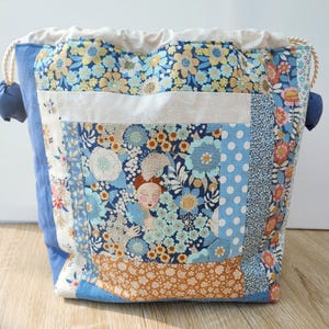Peut inclure: Un sac à cordon fabriqué en tissu patchwork présentant une variété de motifs floraux et à pois dans des tons de bleu, orange et crème. Le sac comprend un panneau central avec une illustration stylisée d'une personne et des liens en corde.
