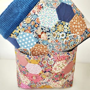 Peut inclure: Un panier de rangement en tissu et une courtepointe assortie avec un motif patchwork. La courtepointe et le panier présentent des motifs floraux dans les tons de rose, bleu, orange et jaune. Le bord du panier est en velours côtelé bleu uni.