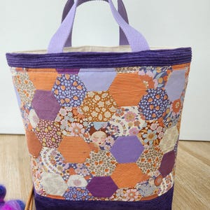 Peut inclure: Un sac fourre-tout composé d'un patchwork de pièces de tissu hexagonales dans des tons orange, violet et crème avec des motifs floraux et rayés. Le sac présente une bordure en velours côtelé violet foncé et des anses en tissu violet clair.