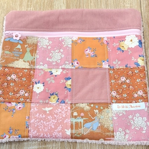 Peut inclure: Une pochette rectangulaire zippée avec un motif patchwork composé de carrés de tissu à fleurs et illustrés dans des tons rose, orange et marron. La partie supérieure est en velours côtelé rose et une étiquette blanche indique Les fils de Florestine.