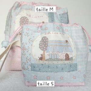 Peut inclure: Deux sacs à cordon de serrage avec un motif patchwork. Les sacs présentent une palette de couleurs bleu clair et rose avec des motifs d'étoiles et de fleurs. Chaque sac a une image centrale d'une maison et d'un arbre, avec le texte "With Love xxx".