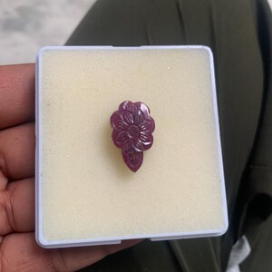 Op de afbeelding: Een gesneden, bloemvormige edelsteen in een kleine, witte plastic doos. De edelsteen heeft een dieprode kleur, met gedetailleerde bloemblad- en bladgravures. De doos wordt in de hand van een persoon gehouden.