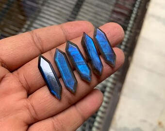 May include: Five iridescent blue labradorite gemstone points, cut into a long, narrow shape, with a pointed tip. The points are arranged in a row, with the largest point at the top and the smallest point at the bottom.