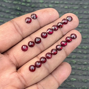 Puede incluir: Una colección de 18 cuentas de piedras preciosas rojas, cada una con una forma redonda facetada. Las cuentas están dispuestas en línea sobre una mano, mostrando su tamaño y brillo.