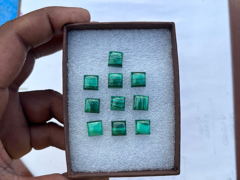May include: A box of twelve green malachite gemstone cabochons, each cut into a square shape. The cabochons are arranged in a grid pattern in the box.