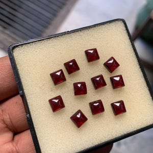 Può includere: Dieci piccole pietre preziose rosso scuro, tagliate a forma quadrata, disposte in gruppo su un inserto in schiuma beige all'interno di una scatola nera. Ogni pietra presenta una superficie sfaccettata che riflette la luce con brillantezza.