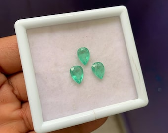 Puede incluir: Tres esmeraldas verdes en forma de pera, exhibidas en una bandeja cuadrada blanca. Las piedras preciosas se colocan sobre un fondo rosa claro, mostrando su claridad y color. La bandeja es sostenida por una mano, con un fondo morado.