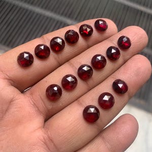 Puede incluir: Una colección de 16 cabujones de piedras preciosas rojas, cada uno con una forma redondeada facetada. Las piedras están dispuestas en filas sobre una mano, mostrando su tamaño y color.