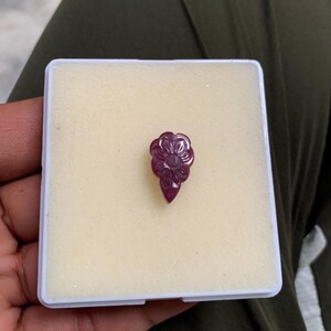 May include: A carved, deep red gemstone in a floral design. The gemstone is set in a white box with a light yellow interior. The gemstone is held in a person's hand, with a dark green background.