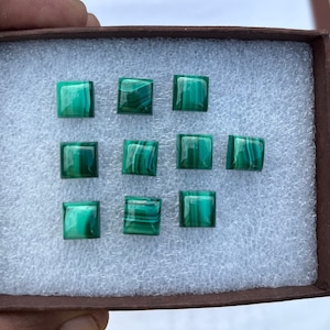 May include: A close-up of a box containing nine square-shaped green malachite gemstones. The gemstones are arranged in three rows of three.
