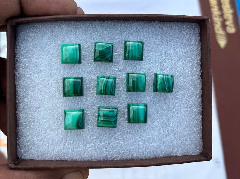 May include: A close-up of a box containing nine square-shaped green malachite gemstones. The gemstones are arranged in three rows of three.