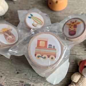 Könnte beinhalten: Eine Sammlung von Holzspielzeugteilen, darunter vier runde Holzmarken mit Tier- und Fahrzeugillustrationen. Die Marken zeigen eine Kuh, eine Giraffe, einen Zug und ein Luftschiff. Weitere Holzspielzeuge und ein Teddybär sind ebenfalls sichtbar.