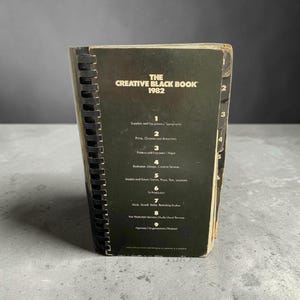 Könnte beinhalten: Ein Vintage-Buch mit Spiralbindung mit dem Titel "The Creative Black Book 1982". Das Cover ist schwarz mit weißem Text und einer Liste von Dienstleistungen. Das Buch ist leicht abgenutzt, mit sichtbaren Seitenkanten und einer schwarzen Spiralbindung.