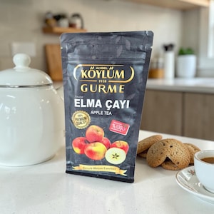 Puede incluir: Un paquete negro de "KÖYLÜM GURME ELMA ÇAYI APPLE TEA" con un grupo de manzanas rojas. El paquete tiene un sello dorado que dice "100% PREMIUM QUALITY" y un sello rojo que dice "%100". También se ven una tetera blanca y galletas.