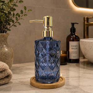 May include: A blue glass soap dispenser with a geometric diamond-cut pattern and a shiny gold-toned pump mechanism. The dispenser sits on a circular light-colored wooden coaster, providing a stable base for the vessel on a countertop surface.