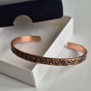 May include: A copper bracelet with an intricate, embossed design. The bracelet is in a white box.