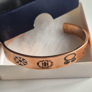 Könnte beinhalten: Ein Kupferarmband mit vier gravierten Symbolen. Die Symbole sind ein Kreis mit einem Punkt in der Mitte, ein Kreis mit einem Kreuz darin, ein Kreis mit einem Stern darin und ein Kreis mit einer Spirale darin.