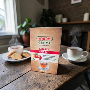 Puede incluir: Una mesa de madera rústica con una caja de té de manzana "KÖYLÜM GURME AMASYA ELMA ÇAYI", con una etiqueta roja y blanca. Dos tazas de té y galletas están sobre la mesa, con vapor saliendo del té. La caja contiene 200 g de té.
