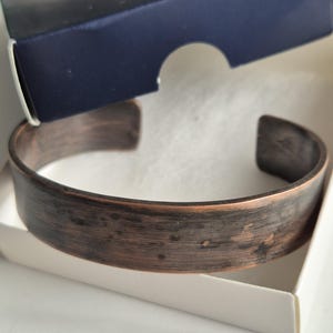 Puede incluir: Un brazalete de cobre marrón oscuro con un acabado martillado. El brazalete está en una caja blanca con una tapa azul.