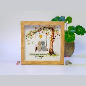 Könnte beinhalten: Ein gerahmtes Kunstwerk mit einem hellen Holzrahmen. Das Kunstwerk zeigt zwei Kieselsteinfiguren auf einer Schaukel unter einem Baum mit einem Herzen. Der Text lautet "Happy 50th Anniversary Stacie & Mike May 26, 1974."