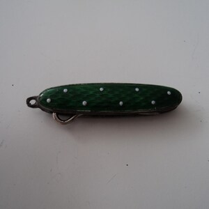 May include: A small, green pocket knife with white dot detailing. The knife features a silver-coloured hook and a small loop at one end. The green surface has a textured pattern. The knife is closed and appears to be a vintage or antique item.