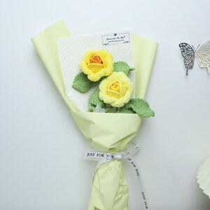 This is a yellow themed knitted rose bouquet containing 2 yellow knitted roses with green cotton thread wrapped around wire to create a rhizome with three leaves on the rhizome. The wrapping paper is yellow and the ribbon is white