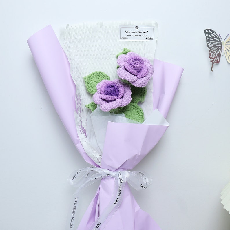 This is a purple themed knitted rose bouquet. It contains 2 purple knitted roses. Green cotton thread is wrapped around wire to create a rhizome with three leaves on the rhizome. The wrapping paper is purple and the ribbon is white