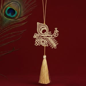 Puede incluir: Un adorno dorado cortado con láser con un diseño de pavo real con una flauta, colgando de un hilo dorado. Una borla dorada cuelga debajo del adorno. Una pluma de pavo real está en el fondo contra un fondo burdeos.