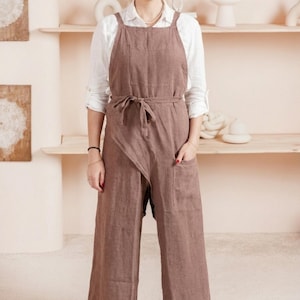 May include: A dusty rose-colored linen apron-style jumpsuit with a cross-back design. The jumpsuit features a tie-waist, large front pockets, and wide legs. The model is wearing a white shirt underneath.