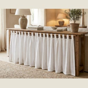 May include: A rectangular wooden console table featuring a natural wood grain finish and a white fabric curtain attached to the front with fabric loops. The table surface holds two stacked books, a ceramic lamp, and a potted green plant.