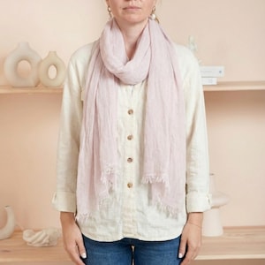 May include: A light pink scarf with a fringed edge is draped around the neck. The scarf is paired with a cream-colored button-down shirt and blue jeans. The background features decorative vases and a shelf.