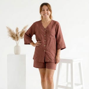 May include: A two-piece set of rust-colored linen clothing. The top is a button-down shirt with a V-neck and three-quarter sleeves. The shorts are high-waisted. The outfit is worn by a person standing in front of a white background.