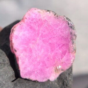 Puede incluir: Primer plano de una gema rosa vibrante y en bruto. La piedra tiene una superficie texturizada con estructuras internas visibles y un borde gris oscuro. La gema se encuentra sobre un fondo gris oscuro.