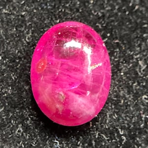 May include: A vibrant, oval-shaped cabochon gemstone in a deep, saturated magenta-pink hue. The surface is smooth and polished, displaying internal crystalline inclusions and natural veining patterns that catch the light with a glossy finish.