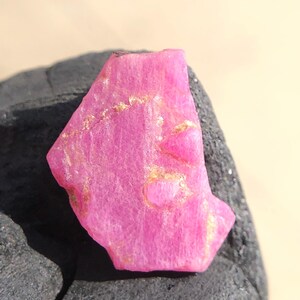 Peut inclure: Une pierre précieuse de rubis rose vif avec des veines dorées. La pierre brute a une surface texturée et est présentée sur un fond sombre et texturé. Cette pierre naturelle est idéale pour la joaillerie ou la collection.