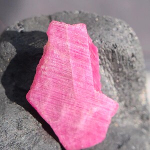 May include: A vibrant pink ruby crystal with a rough, textured surface. The gemstone is displayed against a dark gray background, highlighting its natural form and color. The crystal's facets catch the light, showcasing its internal structure.