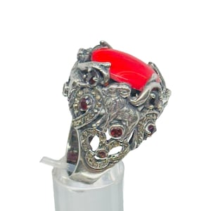 Könnte beinhalten: Ein silberfarbener Ring mit einem großen, ovalen, leuchtend roten Stein als Mittelpunkt. Der Ring hat ein aufwendiges, detailliertes Design mit kleineren roten und klaren Akzentsteinen. Der Ring wird auf einem transparenten Kunststoffständer vor weißem Hintergrund präsentiert.