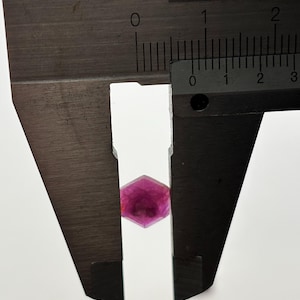 May include: A faceted, hexagonal, deep pink gemstone is measured with a metal caliper. The caliper's scale shows measurements in inches. The gemstone's edges are clearly defined, and the color is consistent throughout.