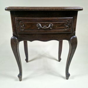 Puede incluir: Una mesita de noche antigua de color marrón oscuro con un solo cajón y un tirador de metal ornamentado. La mesita tiene patas curvas y tallas decorativas en el frente del cajón. La madera tiene una veta visible y un acabado ligeramente envejecido.