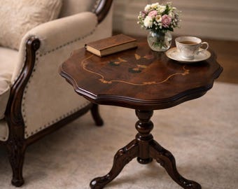 Op de afbeelding: Een donkerbruine bijzettafel in antieke stijl met een geschulpte rand. De tafel heeft een ingelegd ontwerp op het tafelblad, een voetstuk en drie gebogen poten. Een klein boek, een kopje thee en een vaas met bloemen staan op de tafel.