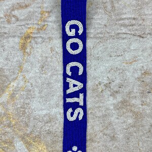 K&ouml;nnte beinhalten: Ein blauer Perlen-Schl&uuml;sselanh&auml;nger mit einem goldenen Verschluss. Der Schl&uuml;sselanh&auml;nger hat wei&szlig;e Perlen, die "GO CATS" buchstabieren, und Pfotenabdr&uuml;cke.