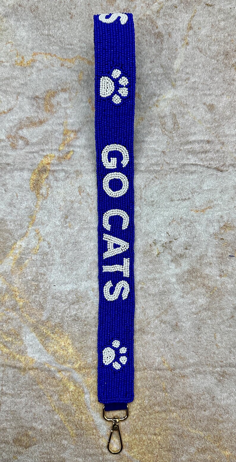 K&ouml;nnte beinhalten: Ein blauer Perlen-Schl&uuml;sselanh&auml;nger mit einem goldenen Verschluss. Der Schl&uuml;sselanh&auml;nger hat wei&szlig;e Perlen, die "GO CATS" buchstabieren, und Pfotenabdr&uuml;cke.