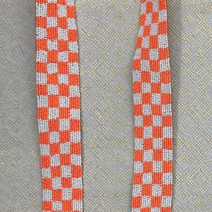 May include: A long orange and white checkered strap with gold metal clasps. The strap is made of woven beads.